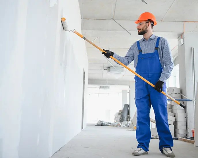 Painter in blue overalls and orange helmet painting a wall white with a roller for basement finishing by Coca Construction.