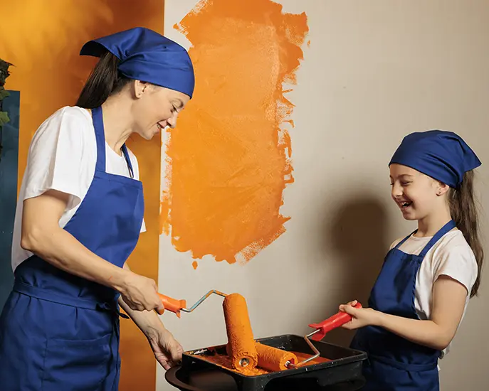 Mother and daughter painting a wall with orange paint, smiling while using paint rollers.