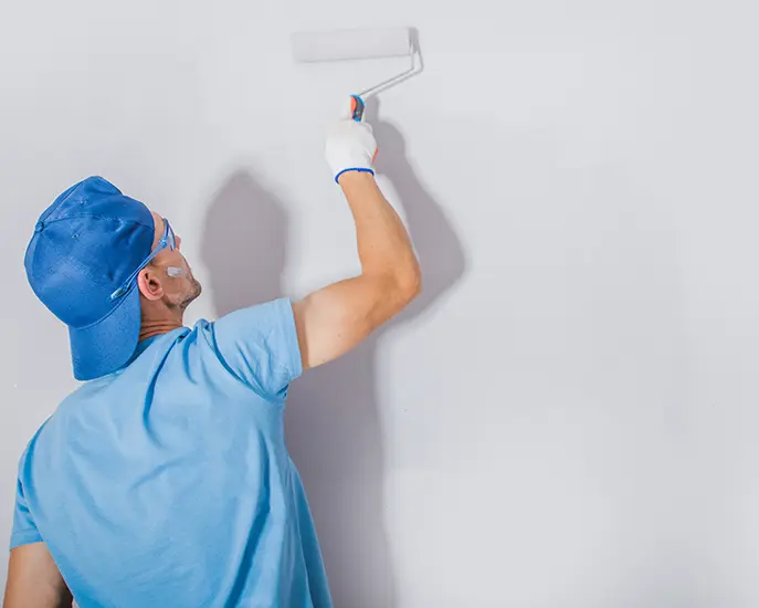 Painter in blue shirt and cap using a roller to paint a residential wall white