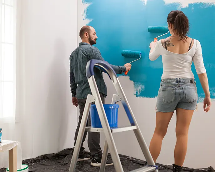 Man and woman painting a wall with blue paint rollers during interior renovation.